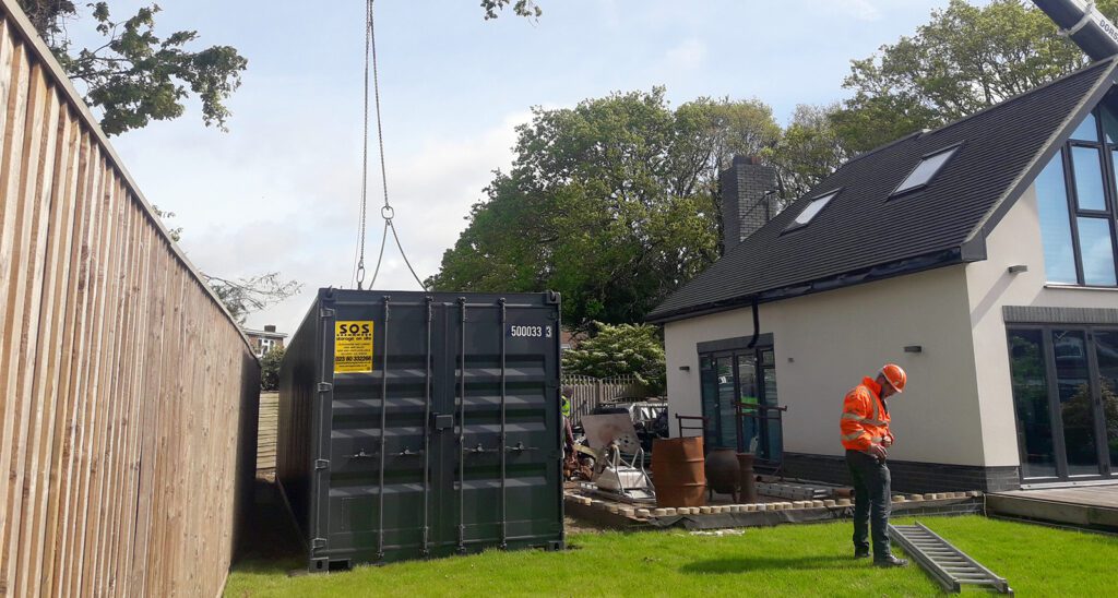 A large shipping container being successfully delivered into a residential back garden via a crane.