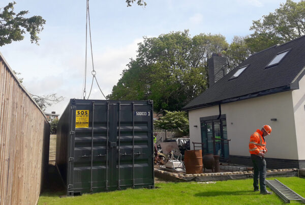 A large shipping container being successfully delivered into a residential back garden via a crane.