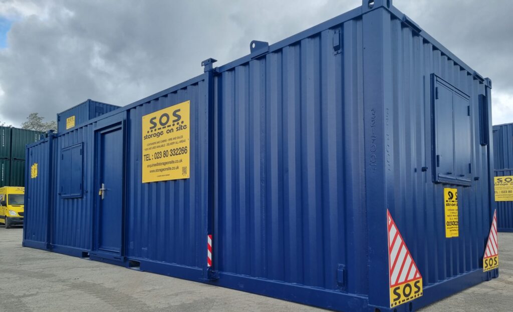Dynamic 3/4 photo of a 32ft blue SOS cabin with yellow advertising board in clean yard surrounded by stacked containers and cabins.