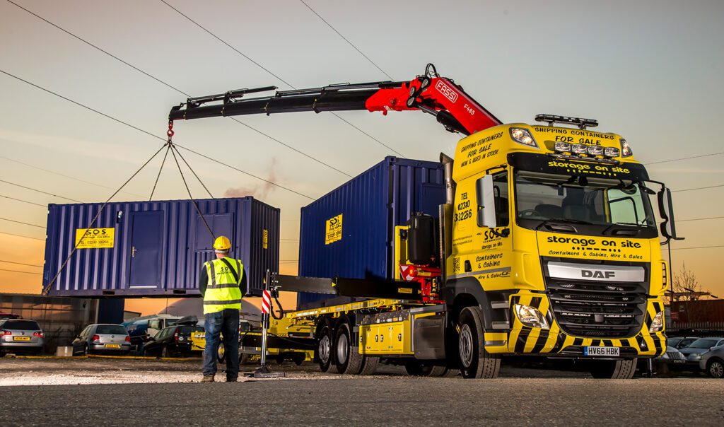 Yellow SOS DAF container delivery truck with FASSI crane offloading a 20ft site cabin at sunset.