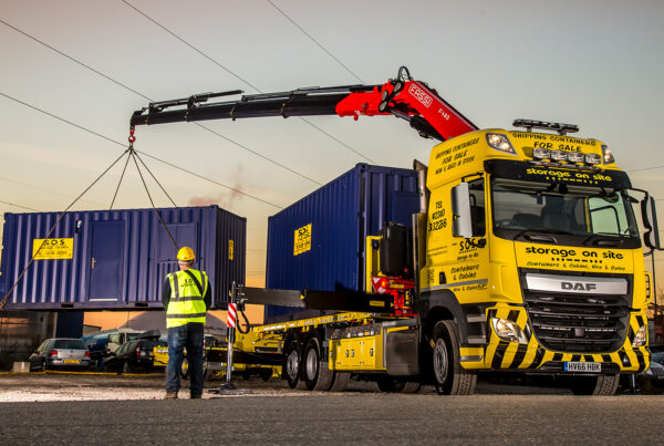 Yellow SOS DAF container delivery truck with FASSI crane offloading a 20ft site cabin at sunset.