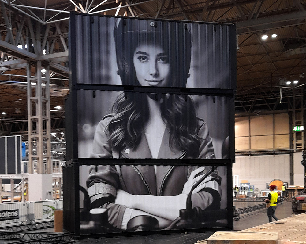 Black shipping containers stacked vertically with a large graphic of a woman wearing a motorcycle helmet, displayed at Motorcycle Live 2024.