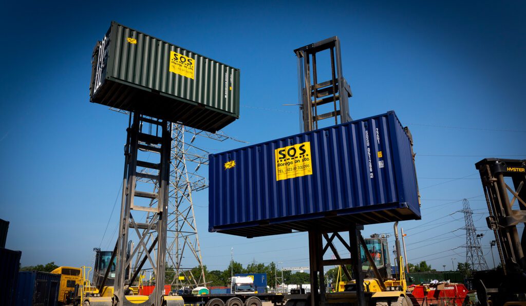 Two Storage On Site shipping containers being lifted by heavy-duty fork lift machinery at the SOS yard under a clear blue sky.